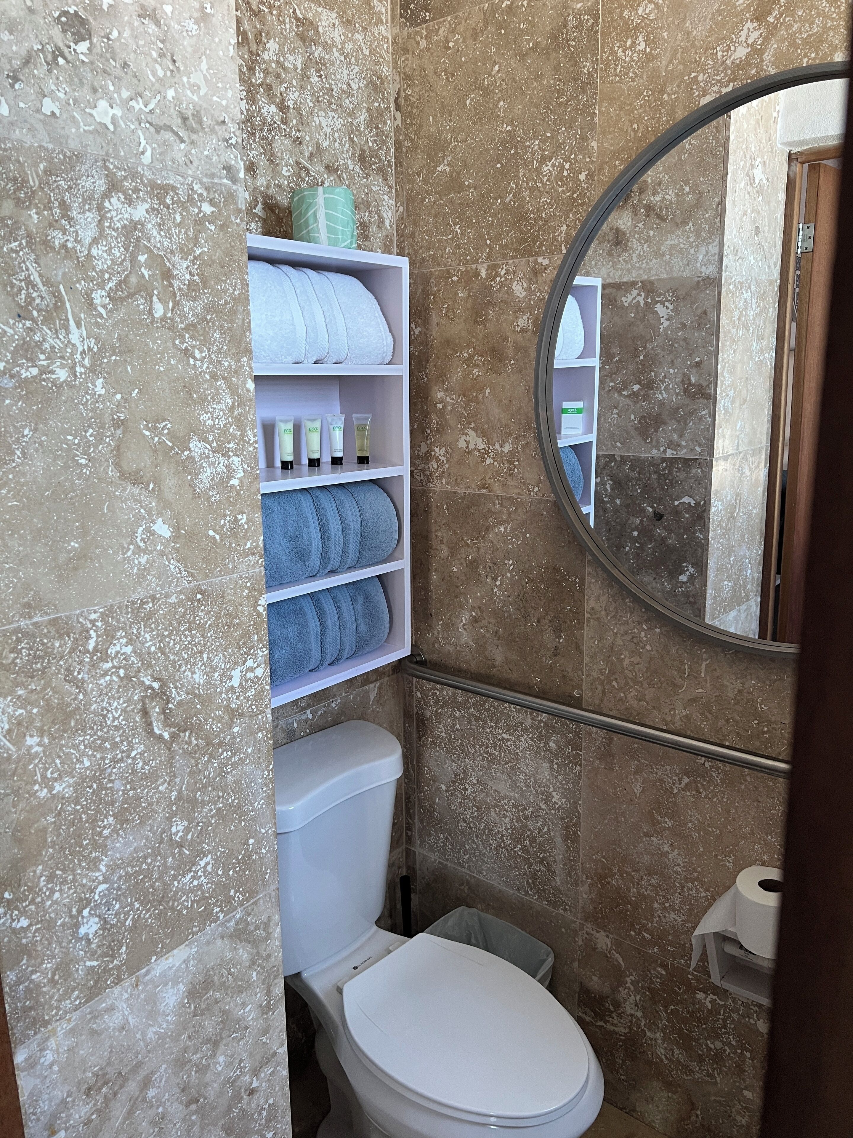 A bathroom with a toilet, a shelf with toiletries, a towel rack, and a mirror.
