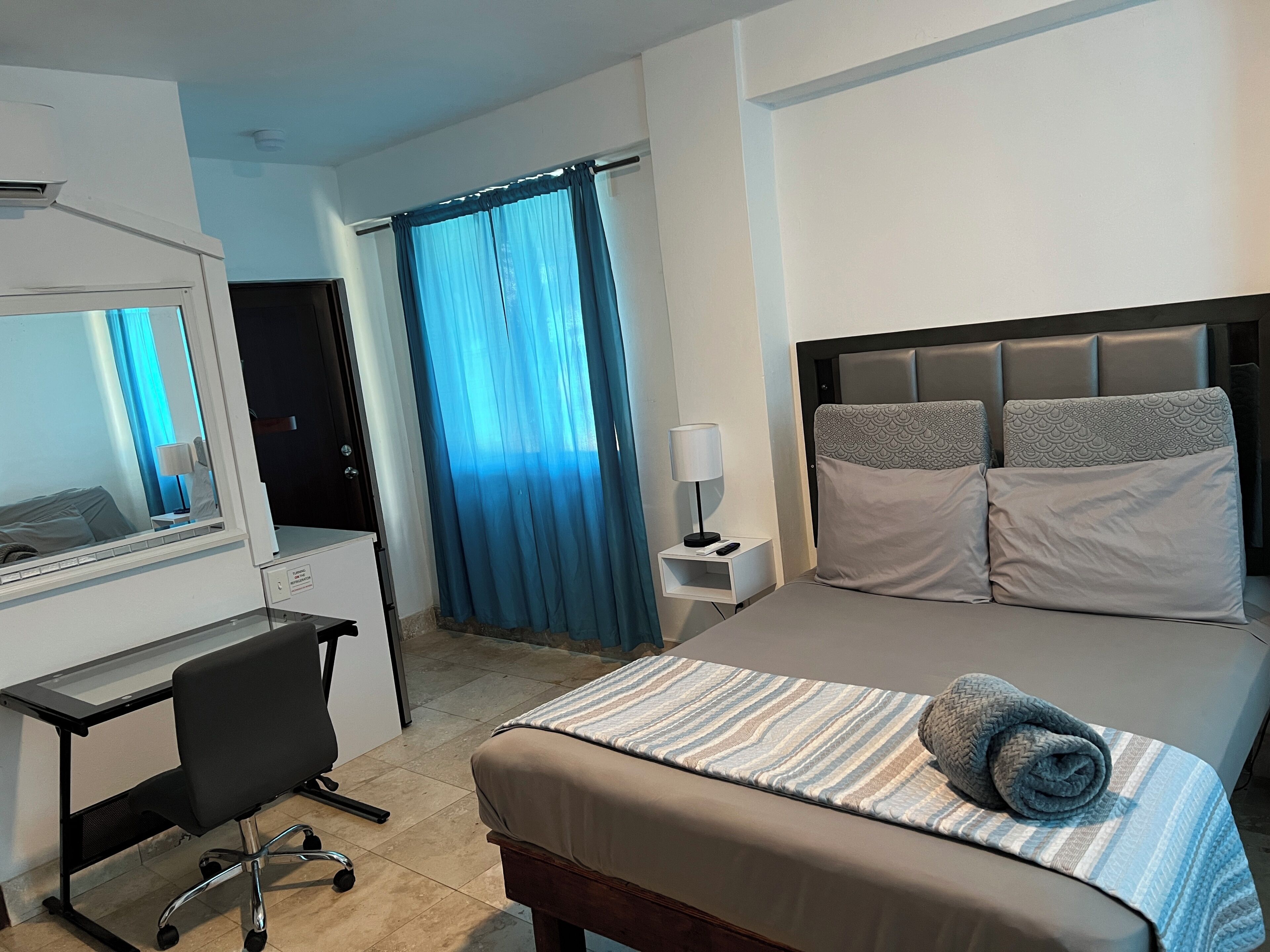 A hotel room with a bed, a desk, a chair, a nightstand, a lamp, and a window with curtains.
