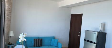 A compact living space with a blue sofa, a glass table with a potted orchid, a ceiling fan, a dark wooden door, and a stainless steel refrigerator.