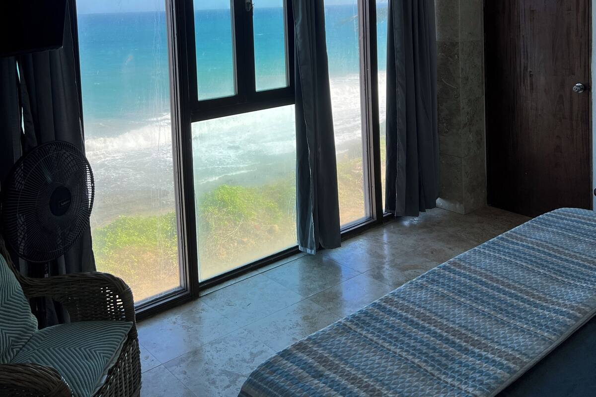 Quarto com cama, ventoinha, guarda-roupa e vista para o oceano através de amplas janelas.