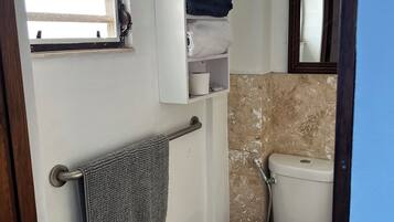 Un baño con inodoro, toallero con toalla, ventana con persianas y estante con toallas y un felpudo enrollado.