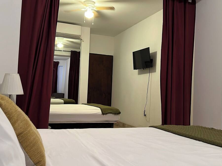 Habitación de hotel con cama, televisor montado en la pared, ventilador de techo y ventana con cortinas.
