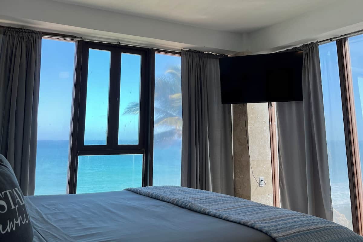 Quarto com cama, ventilador de teto e uma grande janela com vista para o oceano.