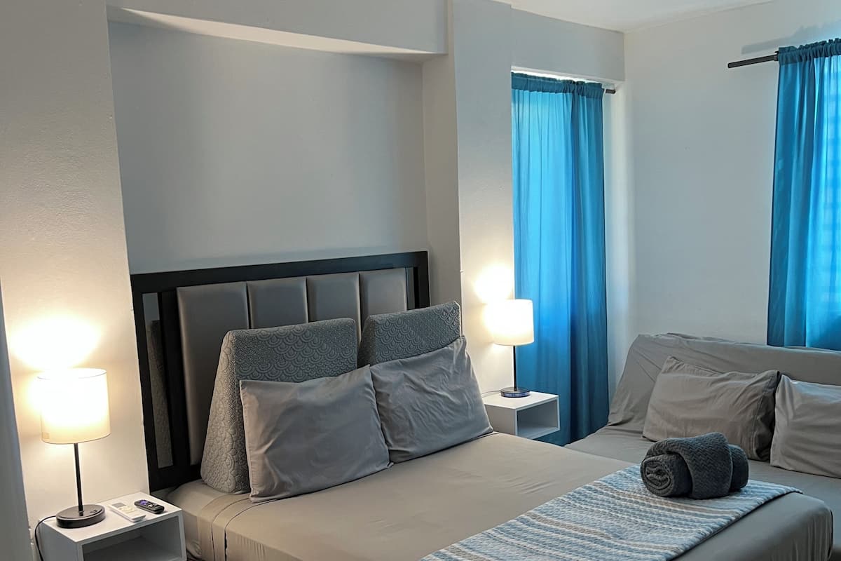 Quarto de hotel com cama, mesas de cabeceira, abajur, cadeira e janela com cortinas.