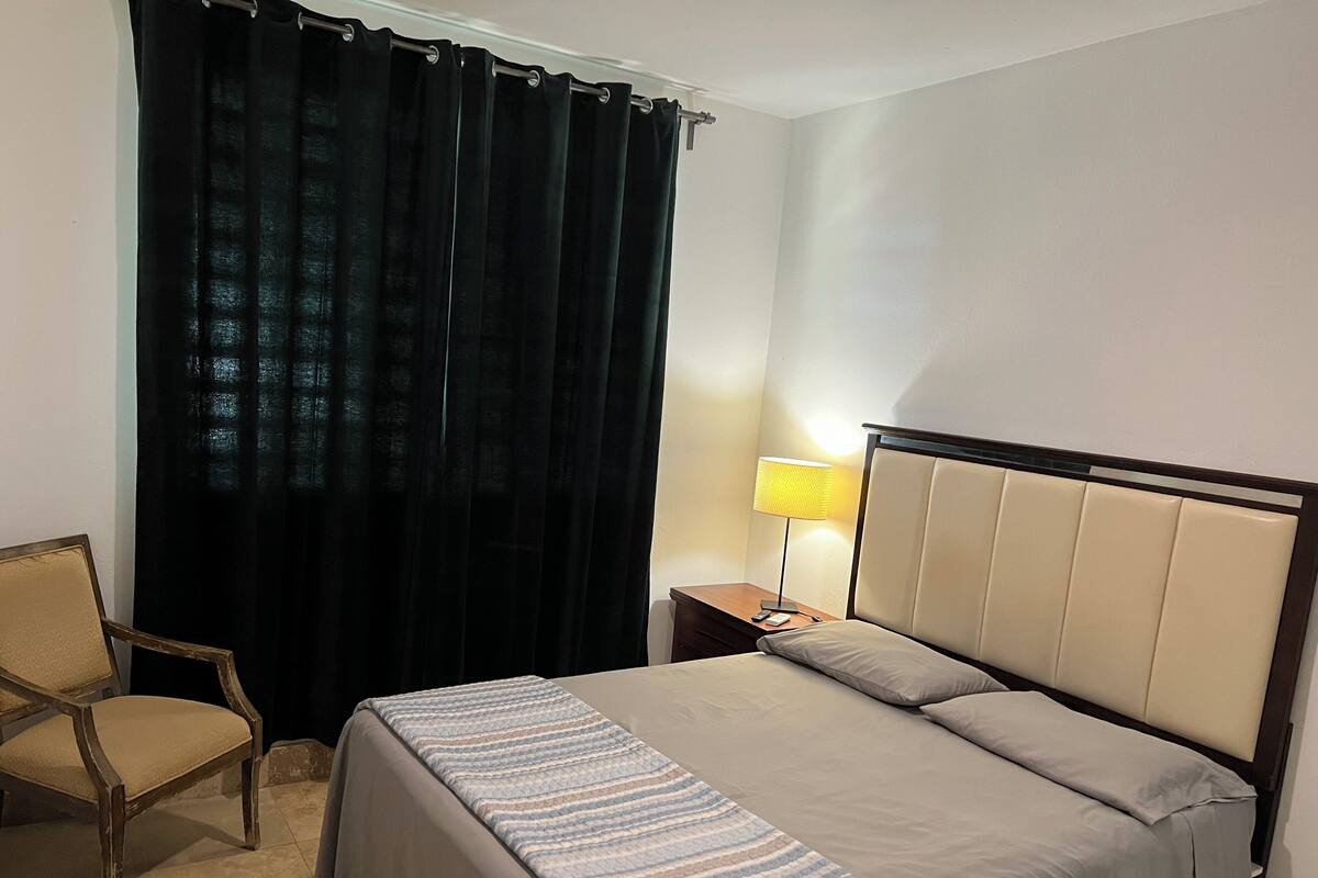 Quarto de hotel com cama, cadeira, mesa de cabeceira com abajur e janela com cortinas escuras.