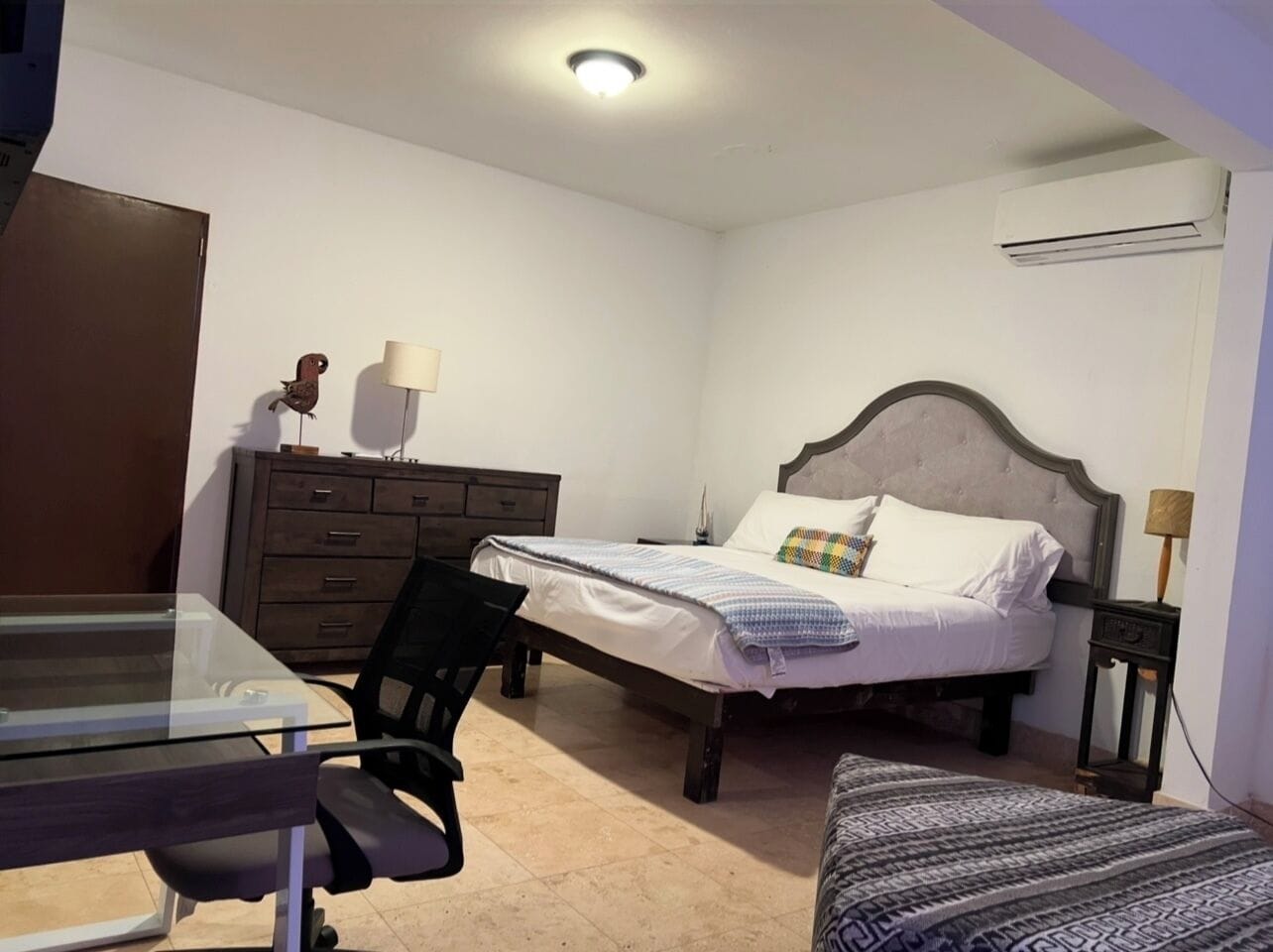 A bedroom with a bed, a desk, a chair, a dresser, and an air conditioning unit.