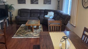 A living room with a dark leather sofa, a wooden coffee table, a dining table with a rug, a floor lamp, and a clock on the wall. - Pondhaven Rereat Fully furnished house with fun and convenience for all ages (Farmington)