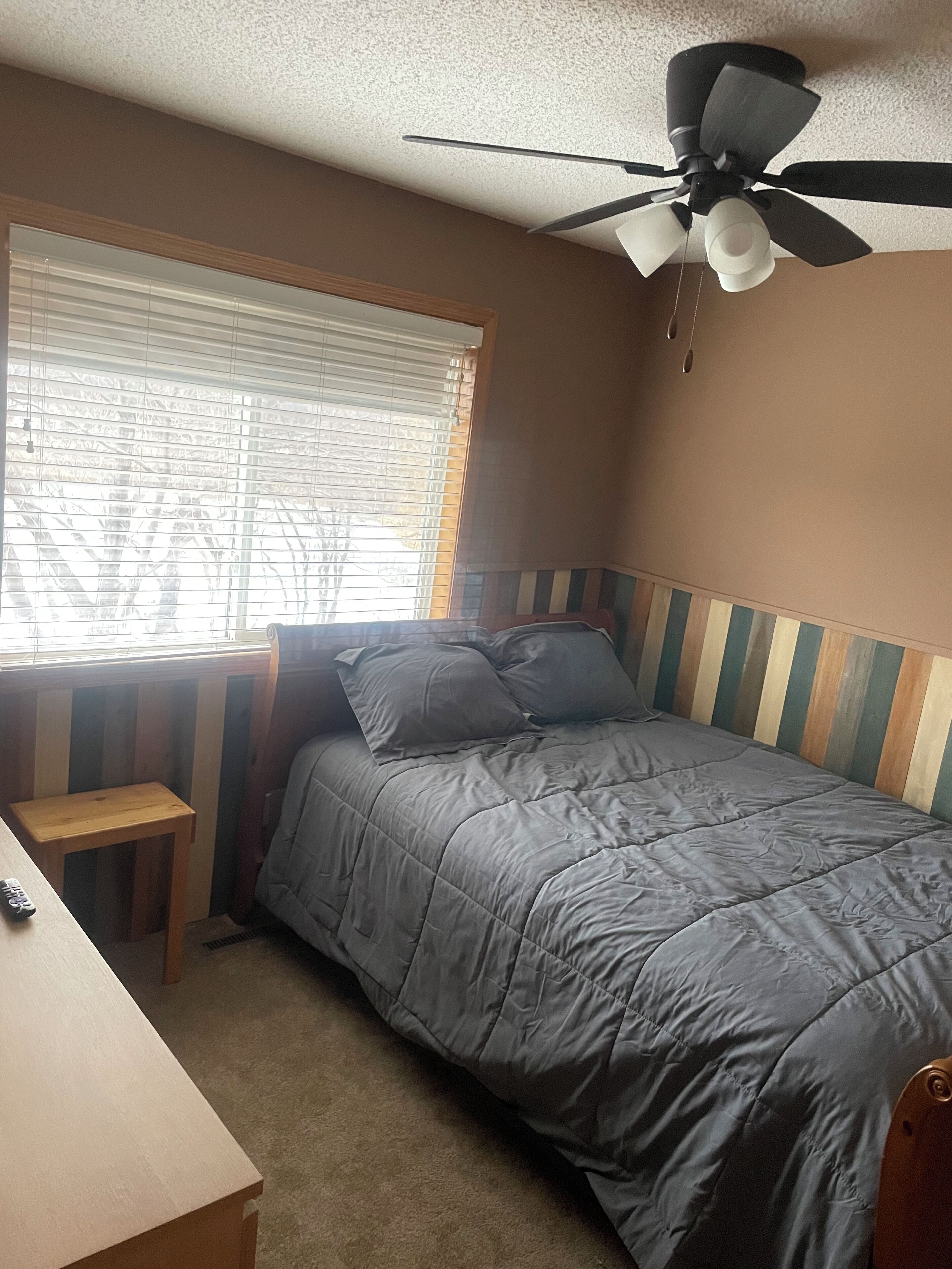 A bedroom with a bed, a desk, a ceiling fan, and a window with blinds.