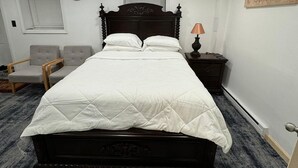 Ein ordentlich eingerichtetes Schlafzimmer mit einem großen Bett, einem Nachttisch mit Lampe, einem Sessel, einer Uhr und einem Fenster mit Jalousien.