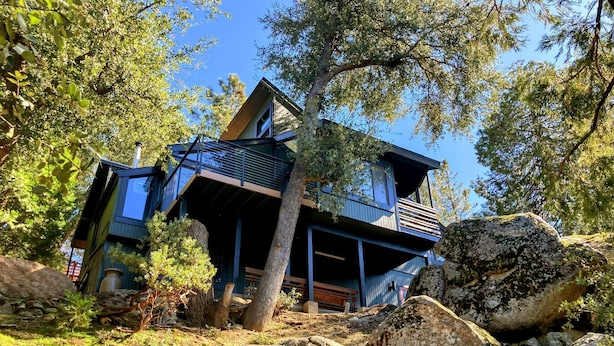 A modern two-story house with a balcony, surrounded by trees and rocks.