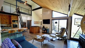 A spacious living area with a high ceiling, a wooden staircase, a kitchen with a bar area, and a large window providing natural light.