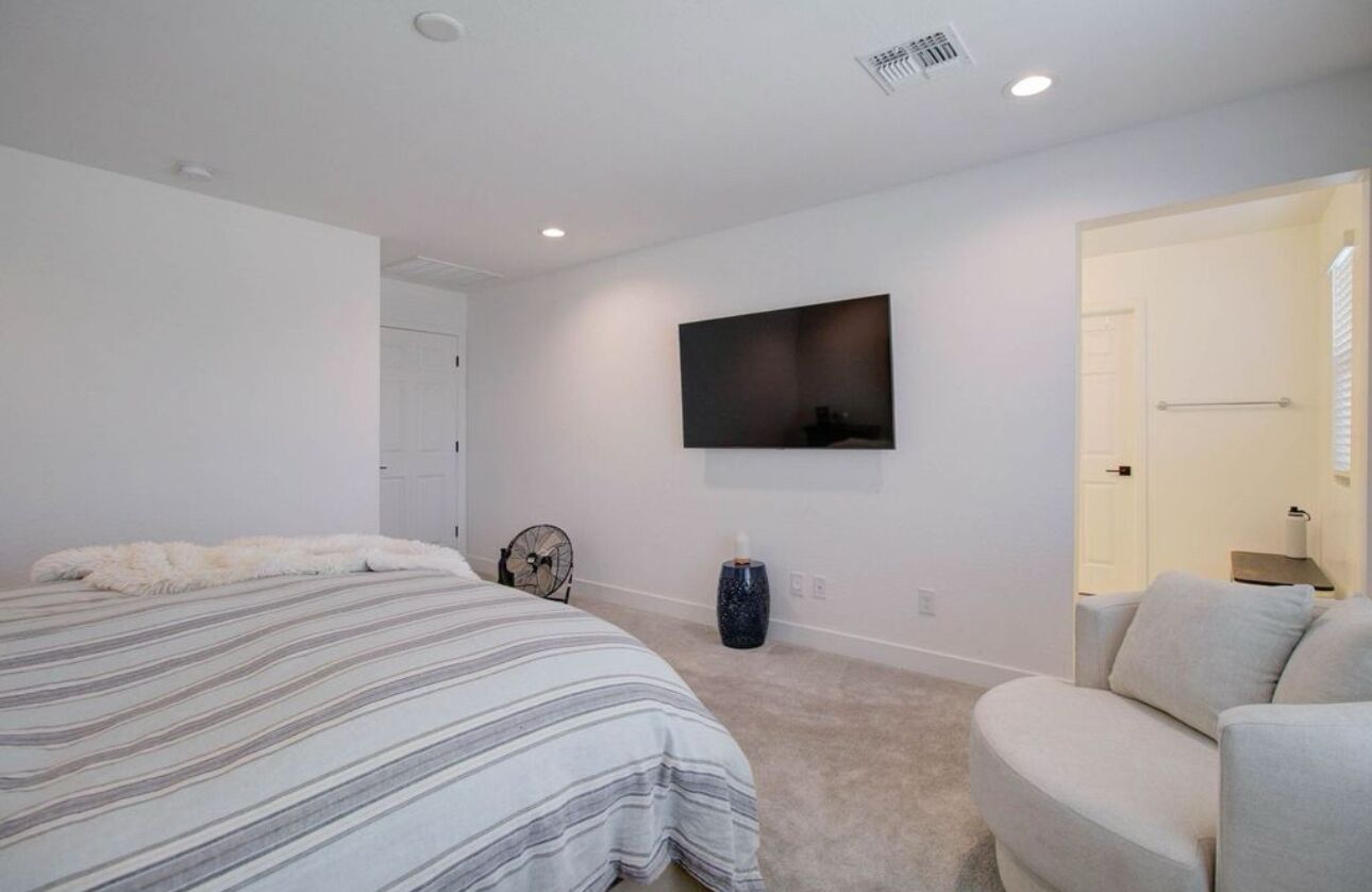 A bedroom with a bed, a sofa, a TV mounted on the wall, and a door leading to another room.