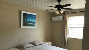 A bedroom with a bed, nightstands, a ceiling fan, an air conditioning unit, a window with curtains, and a framed picture on the wall. - TRINITY BREEZE APARTMENT 1 (Christ Church)