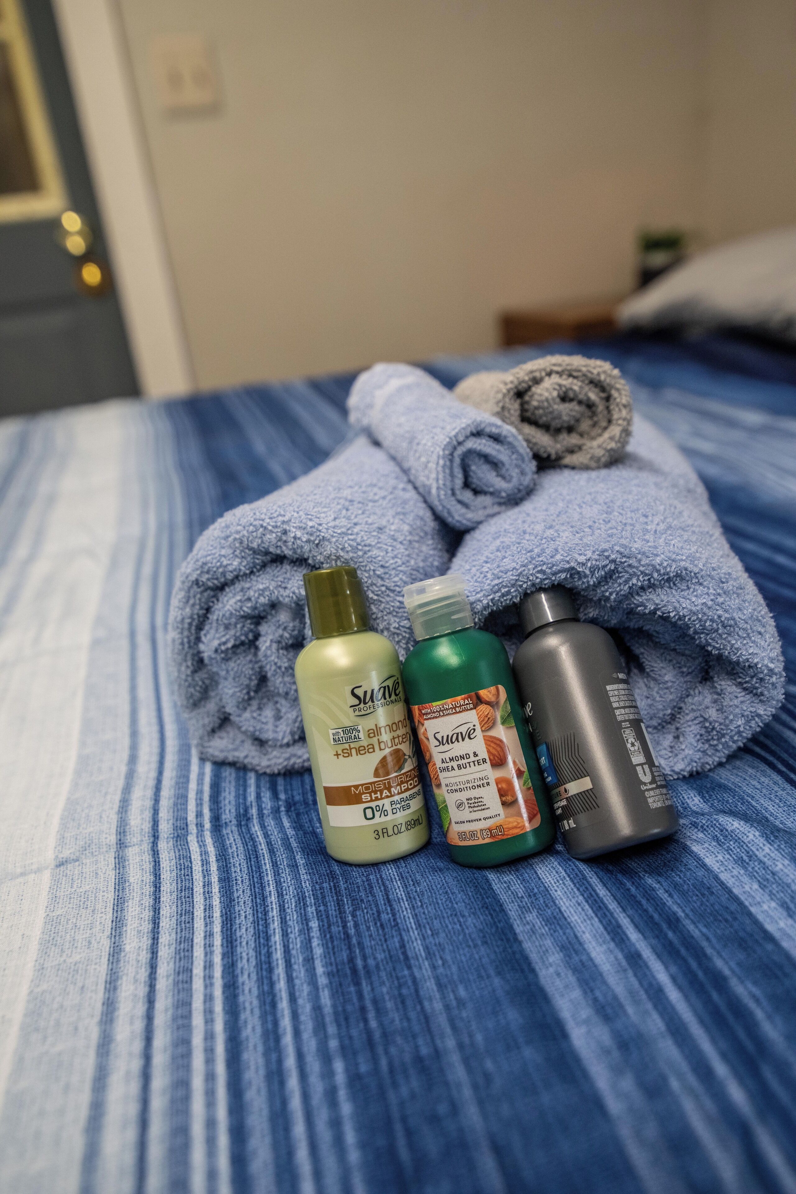 Una cama con una colcha azul a rayas, una toalla enrollada y tres botellas de productos Suave.