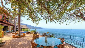 A round glass table with wooden chairs and blue cushions, surrounded by potted plants, overlooking a coastal view with a thatched-roof building and lounge chairs.
