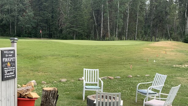 A grassy area with a sign welcoming visitors to a firepit area, surrounded by white chairs and a red trash bin. - Candle Lake retreat backing the 5th green of the golf course. (Candle Lake)