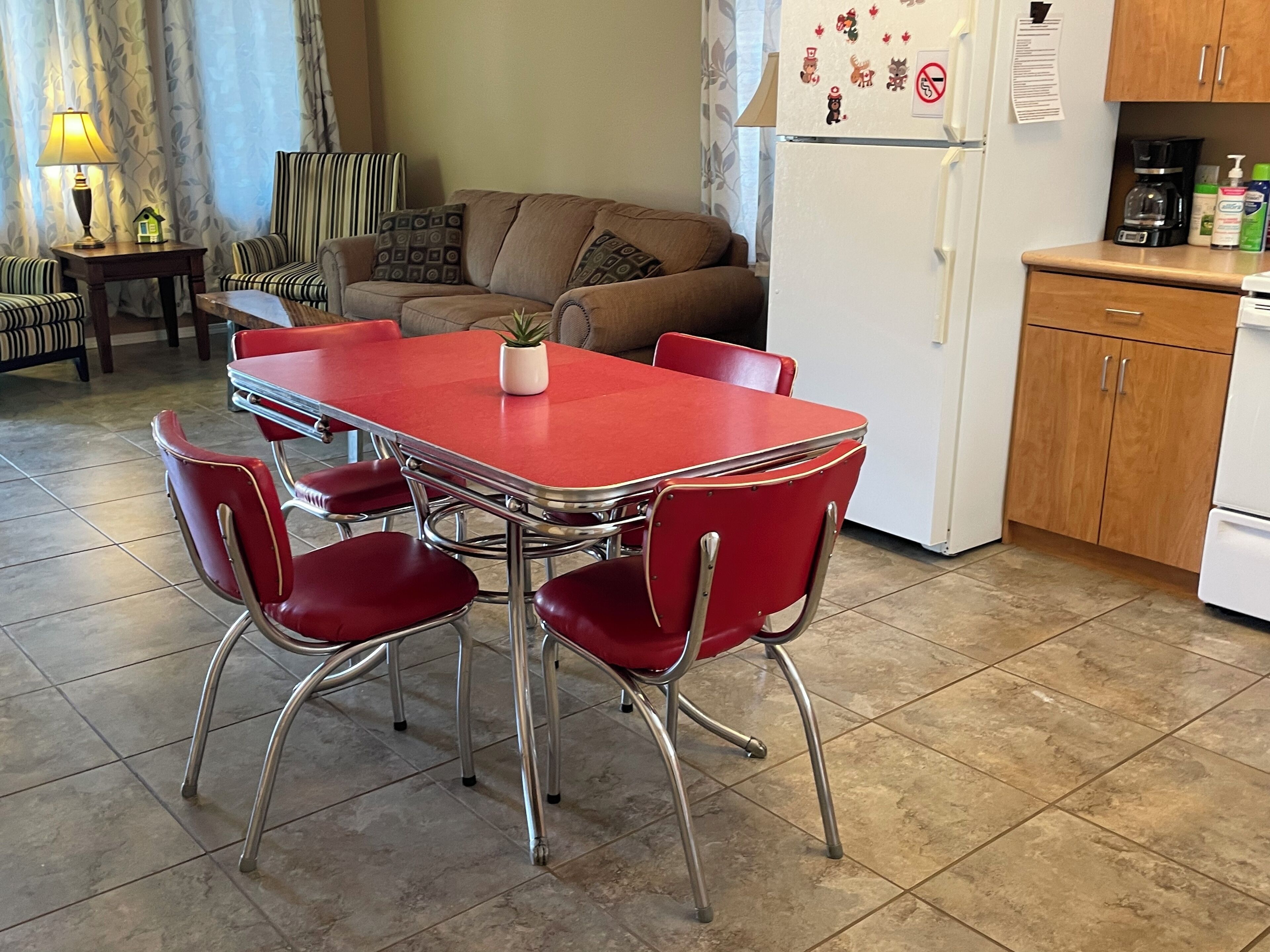 A compact living space with a dining table, red chairs, a sofa, and a kitchen area including a refrigerator and cabinets.