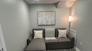 A modern living room with a grey sofa set, a matching ottoman, a patterned rug, and a wall-mounted artwork.