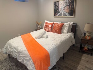 A neatly made bed with a white comforter and an orange throw, a framed cityscape painting above it, and a nightstand with a lamp and a decorative pumpkin.