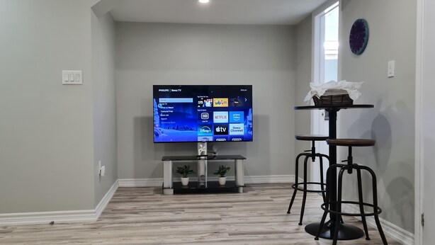 A living room with a flat-screen TV mounted on the wall, a small table with a plant, and a black metal stool.