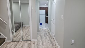 A hallway with a mirrored closet door, a grey sofa, and a kitchen area with a refrigerator and cabinets.