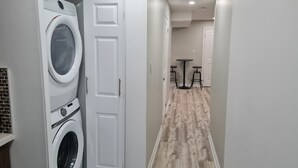 A laundry room with a front-loading washer and dryer, a white door, and a hallway leading to a dining area with a small table and stools.