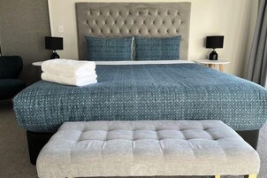 Habitación de hotel con cabecera capitonada, una cama grande con colcha azul estampada, dos almohadas, una mesita de noche con lámpara y un reposapiés gris.