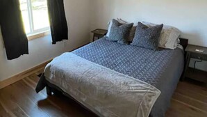 Un dormitorio con cama, suelo de madera, una ventana con cortinas y una mesita de noche.