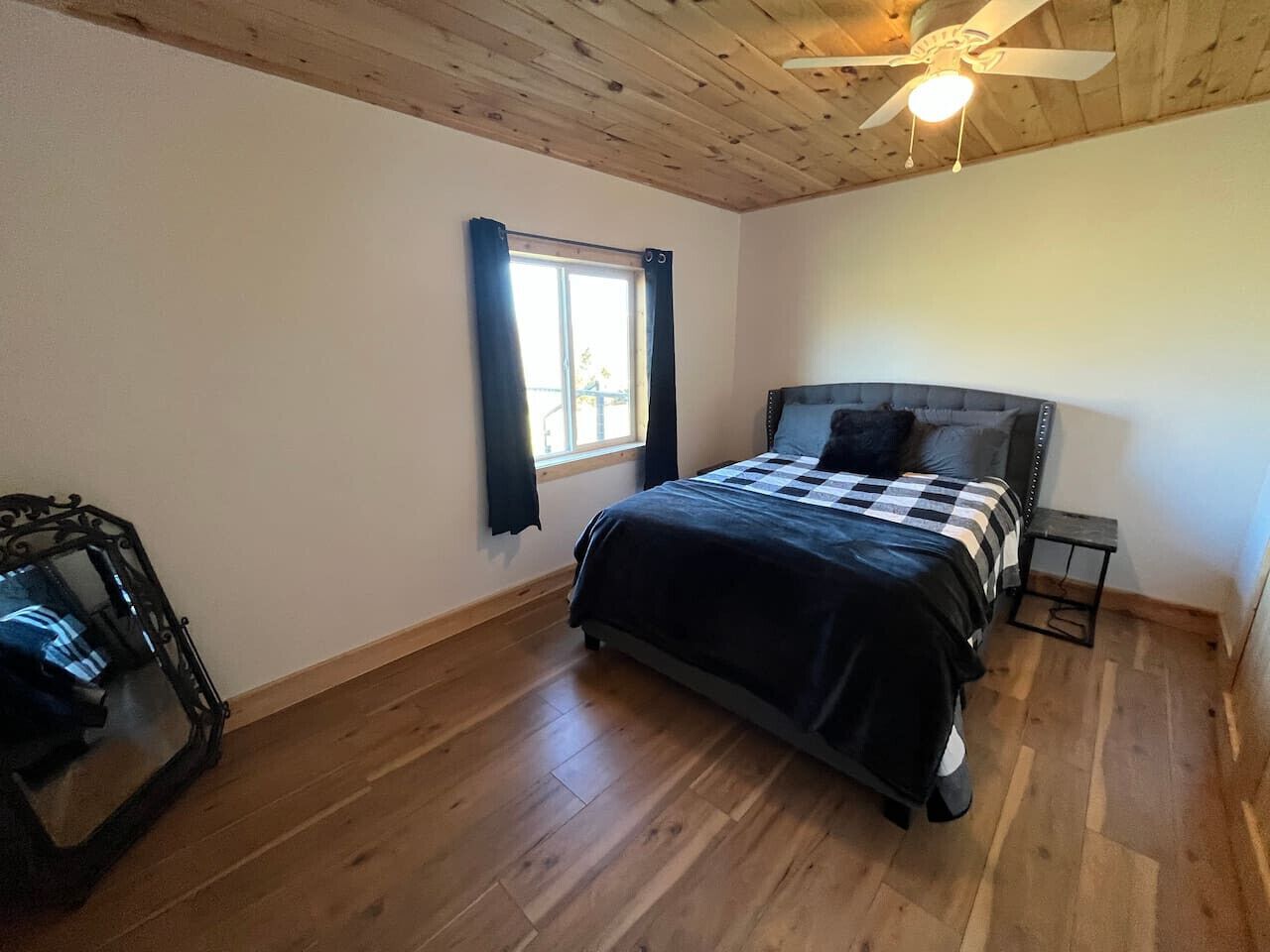 A bedroom with a wooden floor, a bed with a checkered bedspread, a window with curtains, and a ceiling fan.