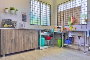 Une cuisine équipée d’un four à micro-ondes, d’un grille-pain et d’une cuisinière à gaz. Des plantes en pot sont disposées sur le mur, et une fenêtre ornée d’un motif à carreaux complète l’ensemble.
