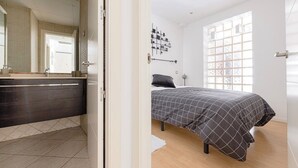 Ein Schlafzimmer mit Bett, Nachttisch, Kommode, einem Fenster mit Jalousien und einer Tür zu einem anderen Raum.