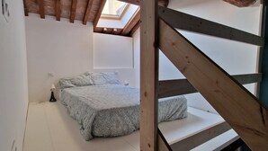 Ein Schlafzimmer mit schrägen Wänden, einem Bett mit gemusterten Betttextilien, einer Nachttischlampe und einem Oberlicht.