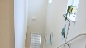 Una escalera con alfombra y barandilla, una ventana con persianas y una pequeña planta en maceta sobre una estantería.