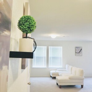 Una sala de estar con un sofá blanco, una planta pequeña en una estantería y dos ventanas con persianas.
