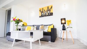 Ein modernes Wohnzimmer mit einem weißen Couchtisch, einem grauen Sofa mit gelben Kissen, einem Bild von Eseln an der Wand und einem kleinen Tisch mit Lampe und Blumen.