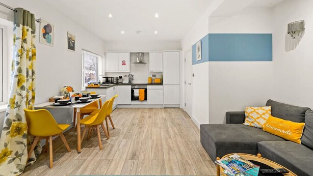 Una cocina moderna con armarios blancos, una zona de comedor con sillas amarillas y un sofá gris con un cojín amarillo.
