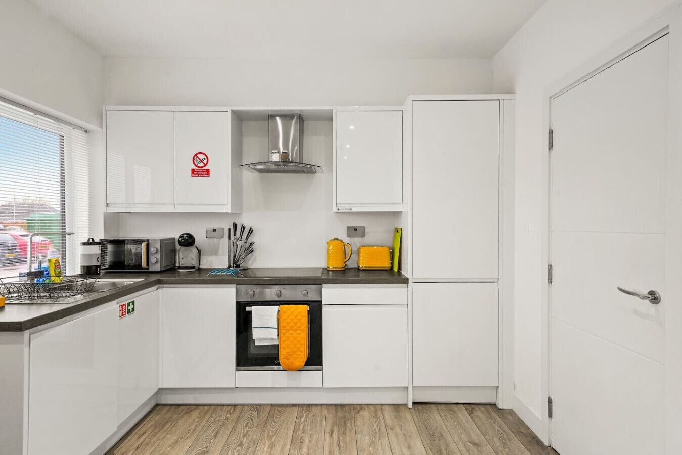 Una cocina moderna con armarios blancos, un extractor de acero inoxidable, horno integrado y refrigerador. Se observa una ventana con persianas, un fregadero con escurreplatos y diversos utensilios y electrodomésticos de cocina.