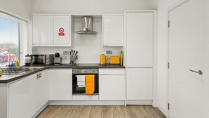 Una cocina moderna con armarios blancos, un extractor de acero inoxidable, horno integrado y refrigerador. Se observa una ventana con persianas, un fregadero con escurreplatos y diversos utensilios y electrodomésticos de cocina.