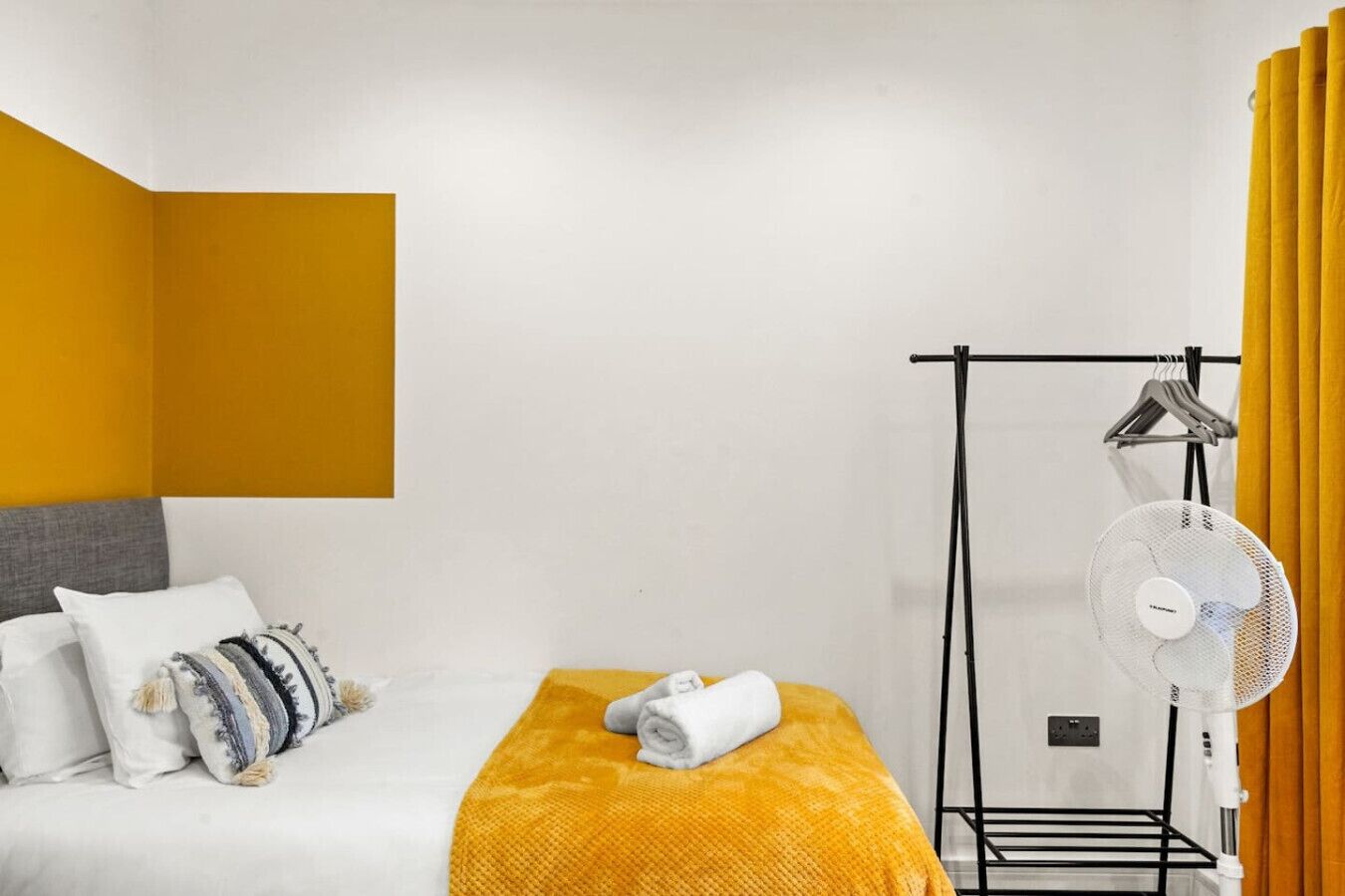A hotel room with a bed, a standing fan, a drying rack, and a yellow curtain.