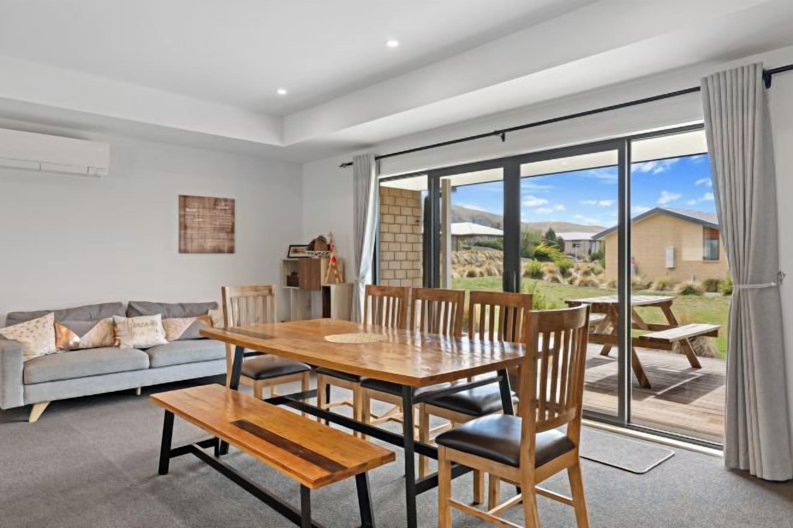 A spacious living room with a large wooden dining table, comfortable seating, and a sliding glass door leading to an outdoor patio.
