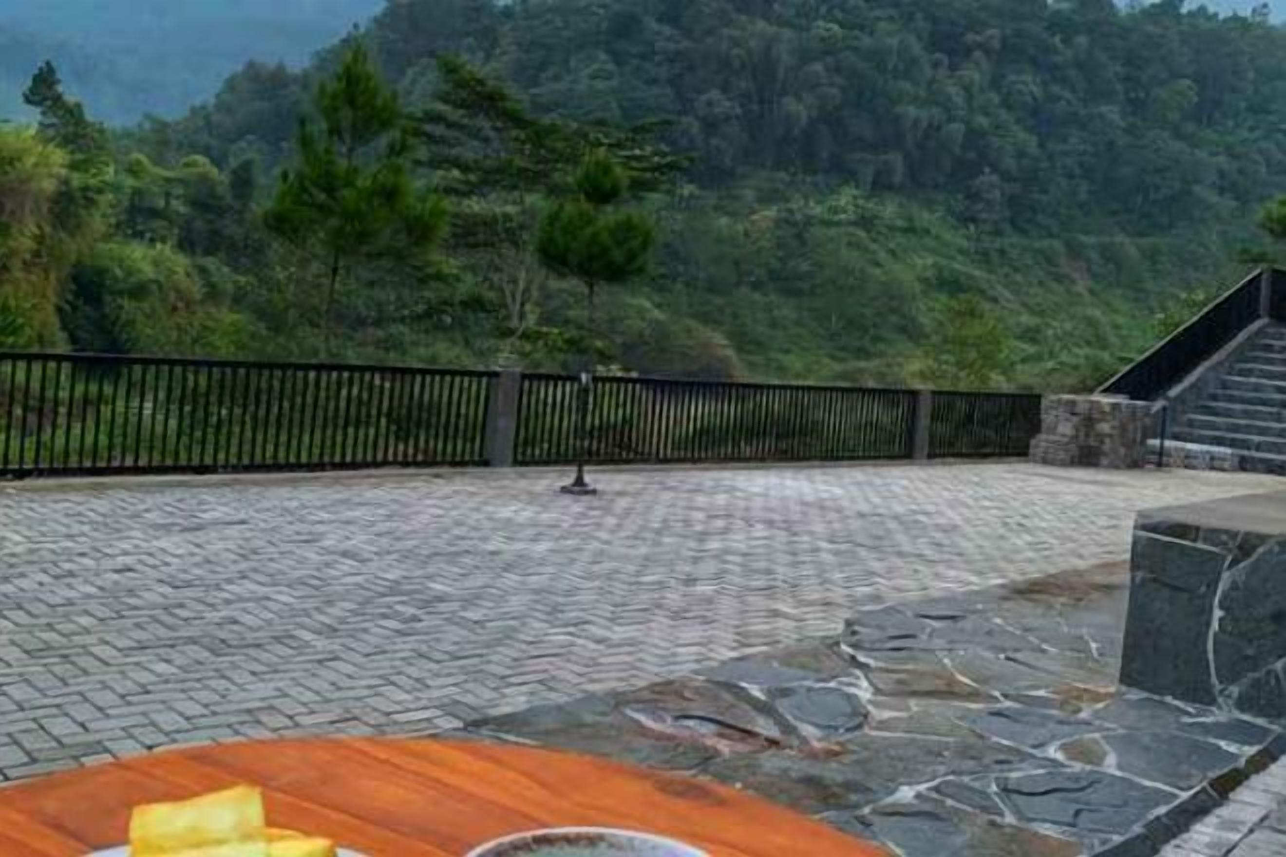 Zona empedrada con un banco de madera y una barandilla de metal, rodeada de follaje y una escalera.