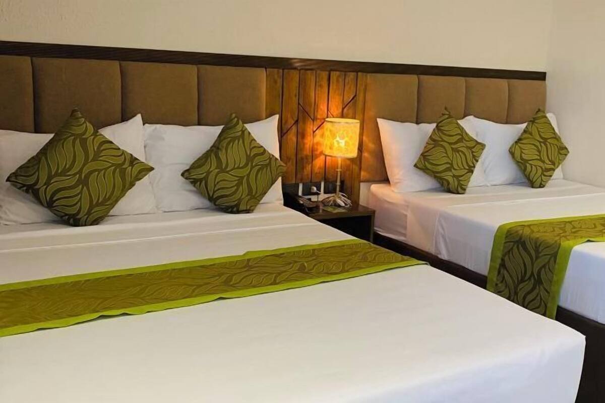 A hotel room with two beds, each with white linens and green leaf-patterned pillows. A nightstand with a lamp is between the beds.