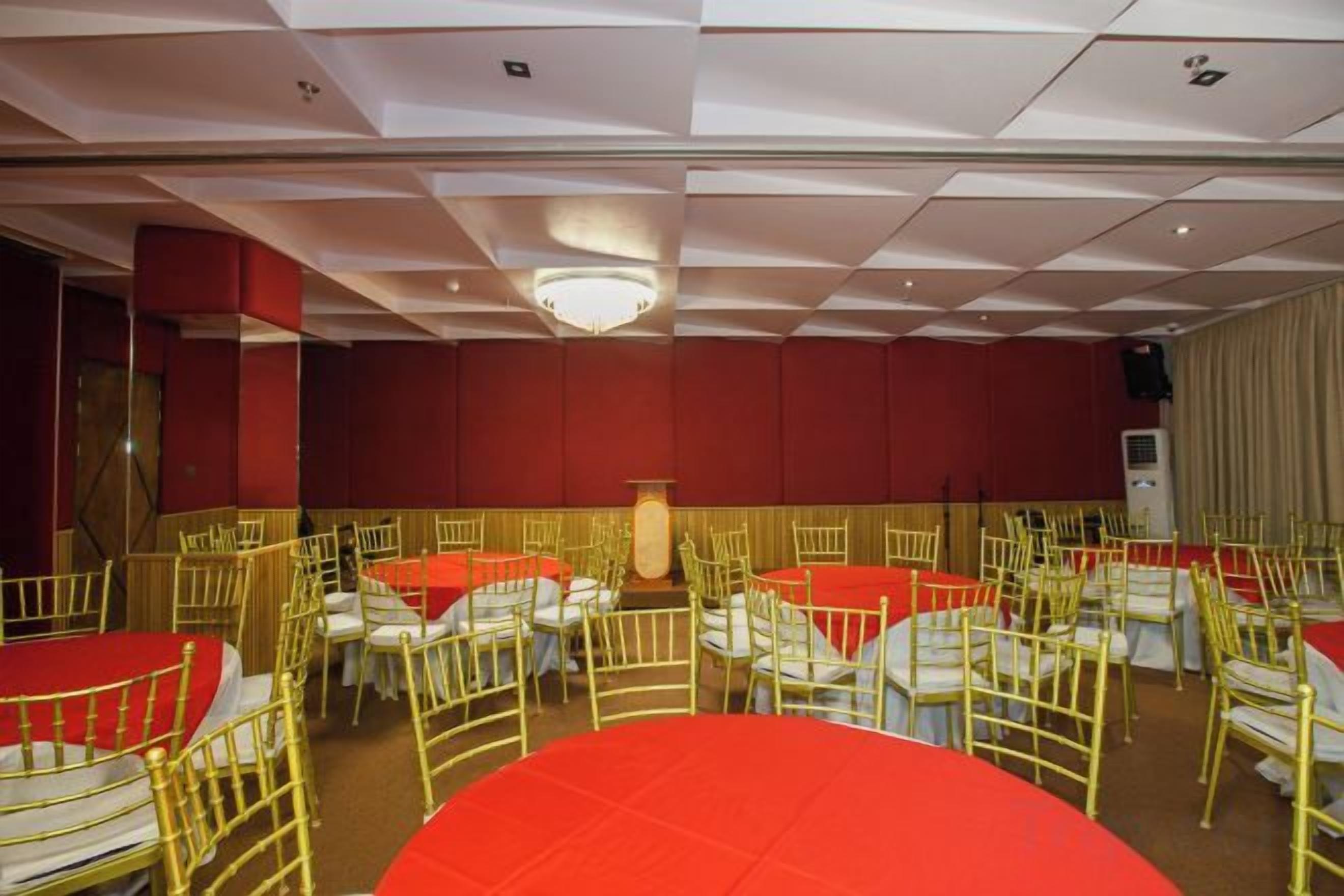 Un salón de banquetes con mesas y sillas redondas, un escenario y una pared roja.