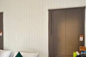 Une chambre d’hôtel avec deux lits, une fenêtre avec des stores et une petite table sur laquelle se trouvent une valise verte et des articles de toilette.