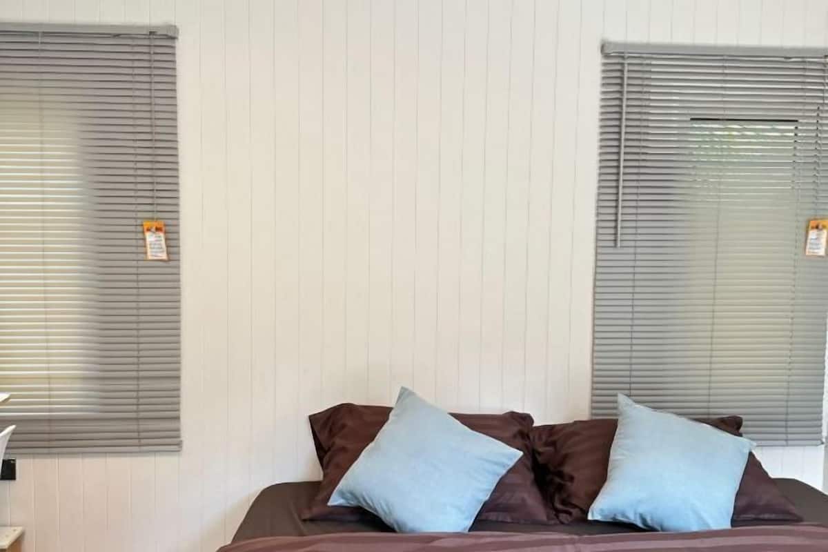 Una cama con almohadas marrones y azules, dos ventanas con persianas y una pared blanca.