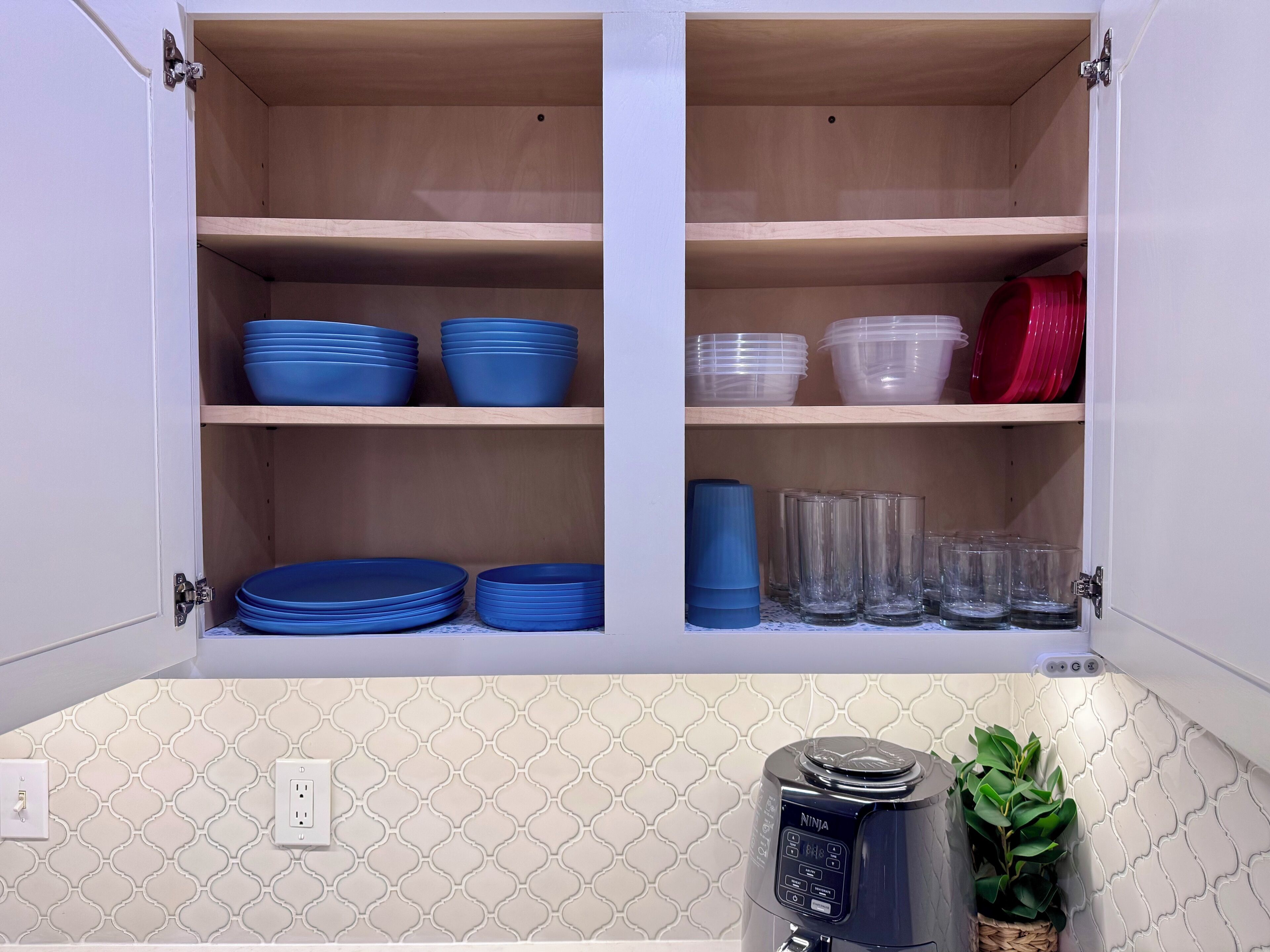 A kitchen cabinet with shelves holding blue and clear plastic dishes, a coffee maker, and a potted plant.