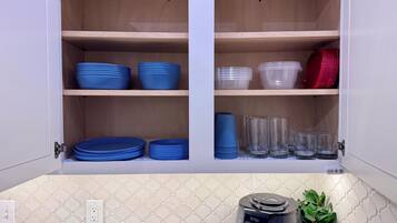 A kitchen cabinet with shelves holding blue and clear plastic dishes, a coffee maker, and a potted plant.