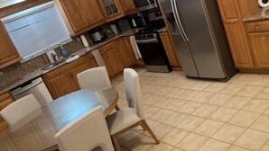 A kitchen with wooden cabinets, a stainless steel refrigerator, a dining table with chairs, and a tiled floor.