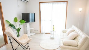 Una sala de estar con un sofá blanco, un sillón color beige, un tapete redondo, una mesita con una botella y un televisor sobre un mueble blanco.