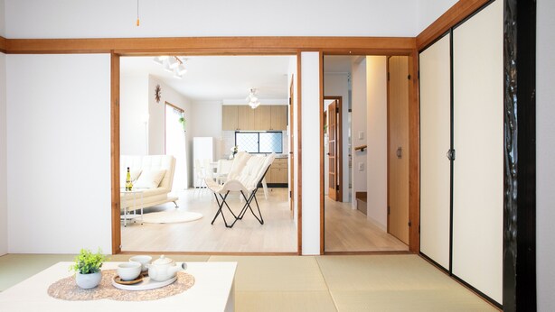 Una habitación tradicional japonesa con suelo de tatami, una mesa baja con juego de té y puertas correderas que dan a una sala de estar con sofá y televisor.